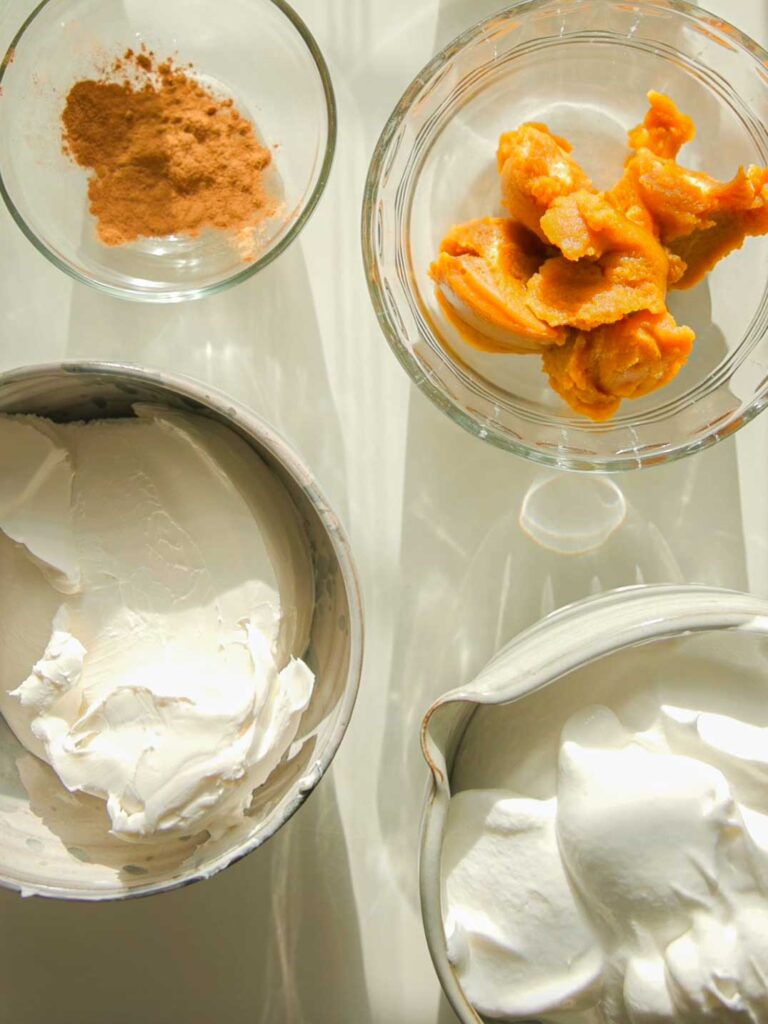 Four bowls on a light table containing the ingredients for this vegan no-bake pumpkin cheesecake: one with cinnamon powder, another with pumpkin puree, a third with whipped cream, and the last with cream cheese. The sunlight casts soft shadows over the ingredients.